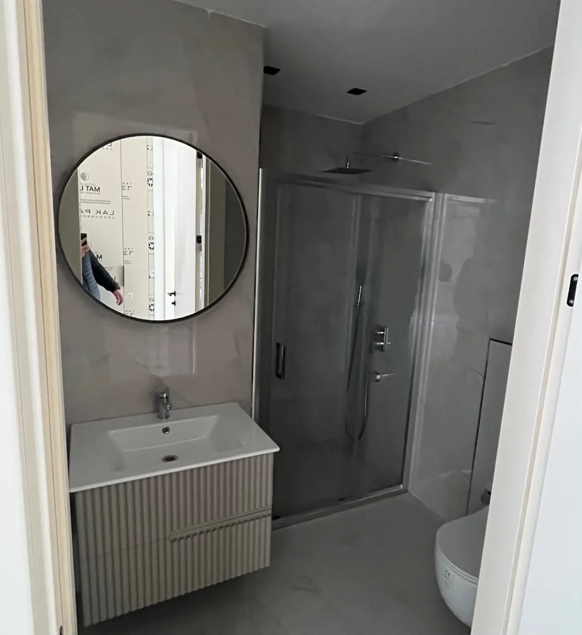 Tepebaşı Batıkent Mahallesi'nde yenilenen ikinci banyo: sürgülü cam duş kapısı, oluklu ön yüzlü lavabo dolabı ve siyah çerçeveli yuvarlak ayna, 2026