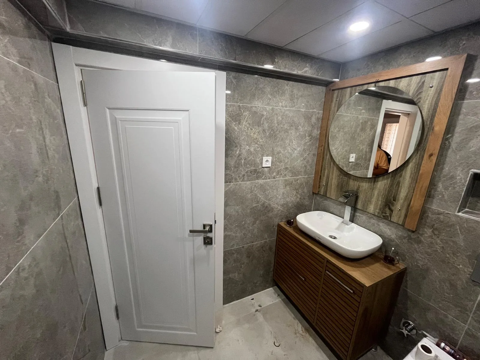 Odunpazarı Gökmeydan Mahallesi'nde yenilenen banyonun kapı tarafı: beyaz panelli kapı, gri karolar, yuvarlak aynalı lavabo dolabı ve duvar nişi, 2026