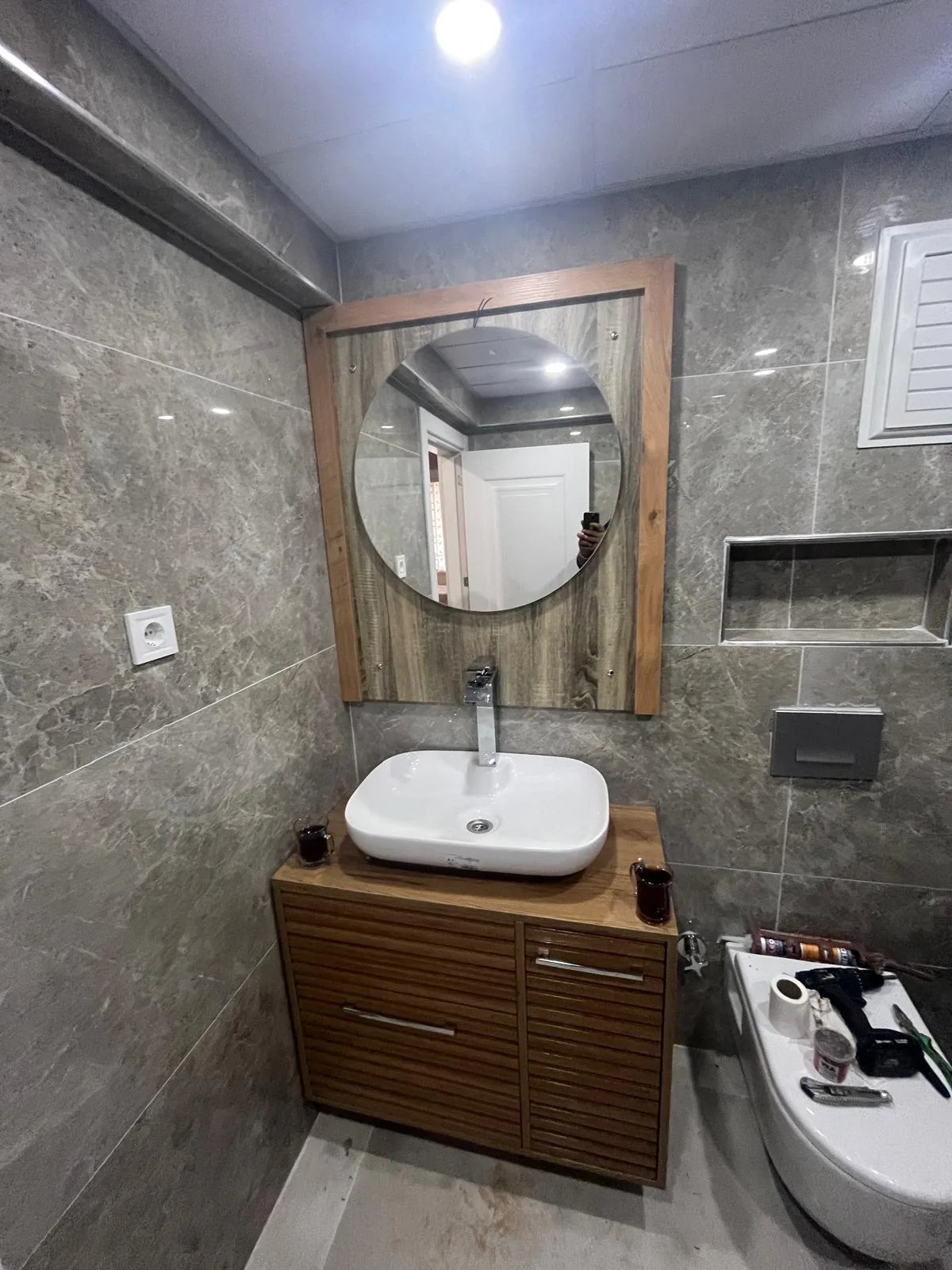 Odunpazarı Gökmeydan Mahallesi'nde son aşamadaki banyo: lavabo dolabı, yuvarlak ayna, duvar nişi, gri kumanda panelli asma klozet ve üzerinde el aletleri, 2026