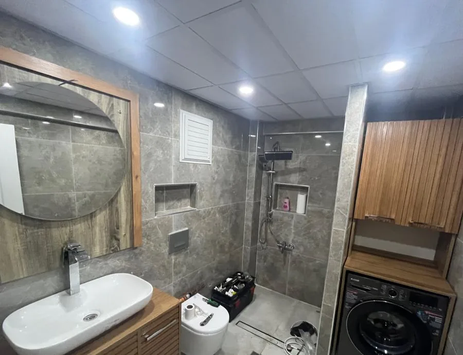 Odunpazarı Gökmeydan Mahallesi'nde yenilenen banyo: gri mermer görünümlü karolar, yuvarlak aynalı lavabo dolabı, asma klozet, duş nişi ve çamaşır makinesi dolabı, 2026