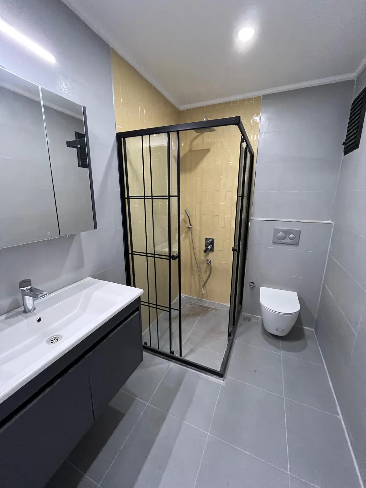 Odunpazarı Sümer Mahallesi'nde yenilenen banyo: gri karolar, sarı tonlu karolu duş alanında siyah profilli ızgara desenli duşakabin, asma klozet ve antrasit lavabo dolabı, 2026