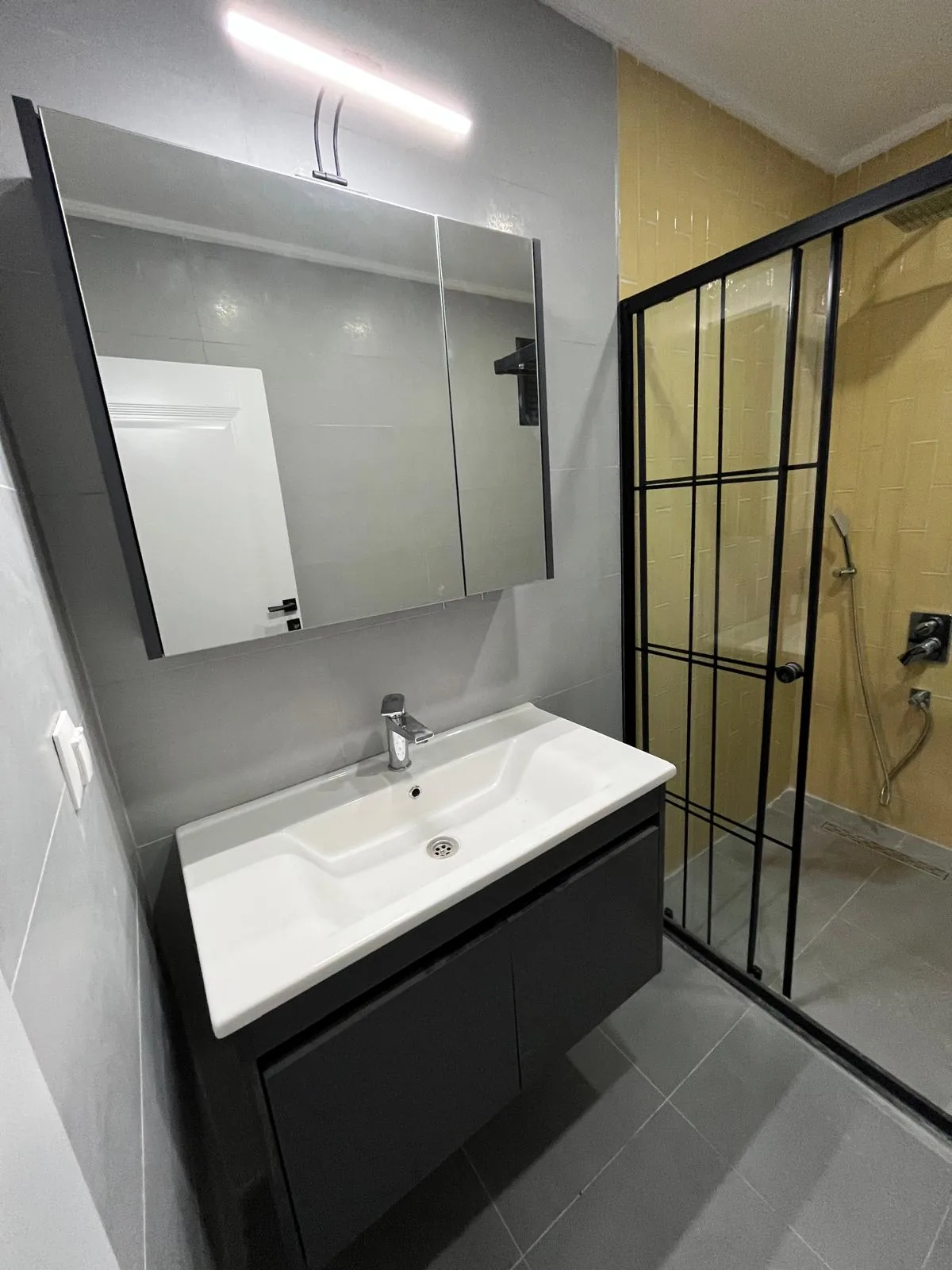 Odunpazarı Sümer Mahallesi'nde yenilenen banyonun lavabo tarafı: antrasit asma dolap üzerinde beyaz lavabo, üstünde aydınlatmalı aynalı dolap, yanında siyah profilli duşakabin, 2026